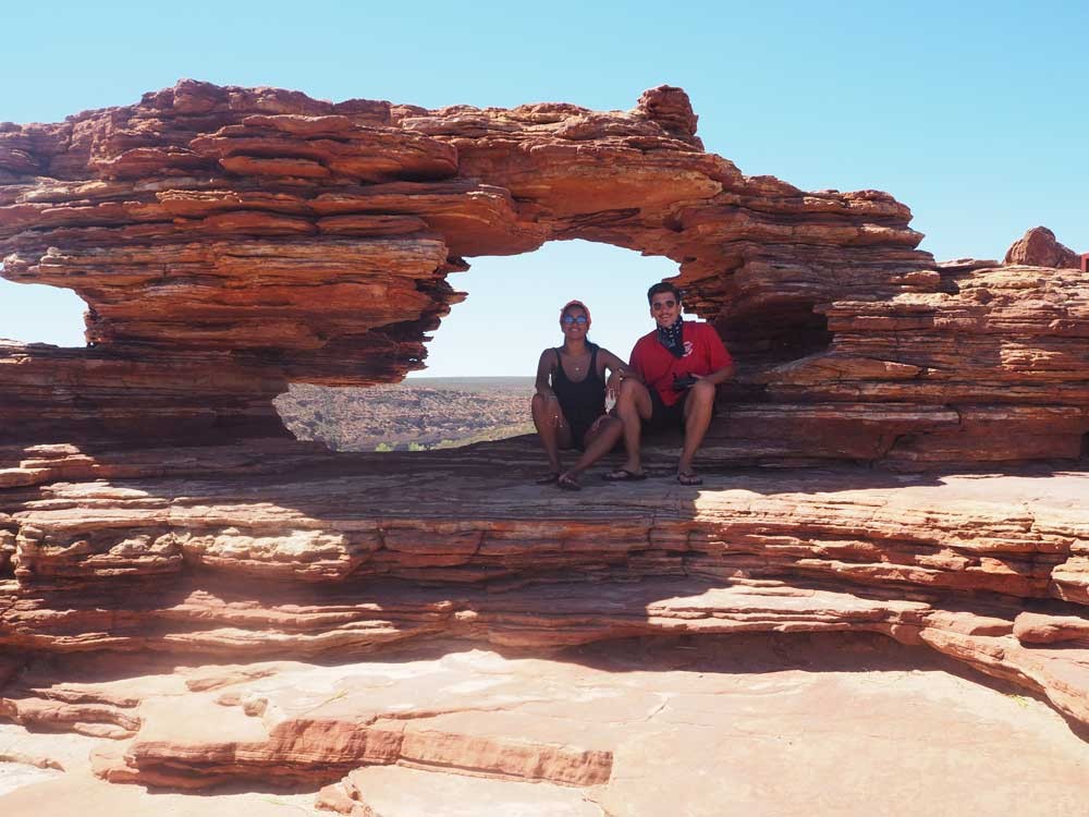 pareja sonriente mirando a cámara y están sentados en unas rocas y detrás el cielo despejado